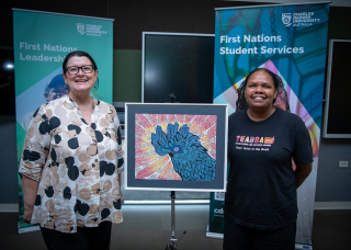Caucasian woman wearing glasses with a proud smile on her face standing next to an artwork of a blue cockatoo with a young Aboriginal woman on the right also with a big proud smile on her face.