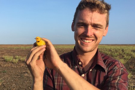 Dr Robin Leppitt head and shoulders, holding a small yellow bird with both hands, green flat plain in background