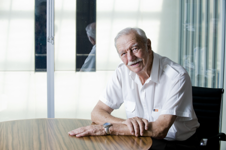 Image of the Honourable Austin Asche AC KC in 2011 sitting at a desk looking straight at the camera. He wears a white shirt with a badge of the Northern Territory flag. 