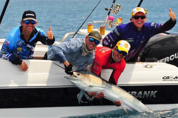 Four men in a boat, two leaning over the side holding a big fish