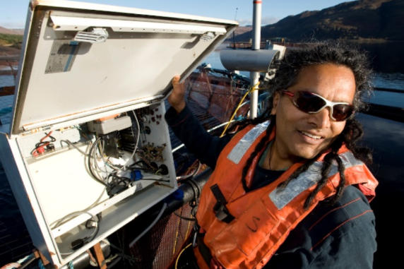 Sunil Kadri, wearing orange life vest, holding up the cover of a vertically mounted box containing electrical circuits, with water and hills in the background