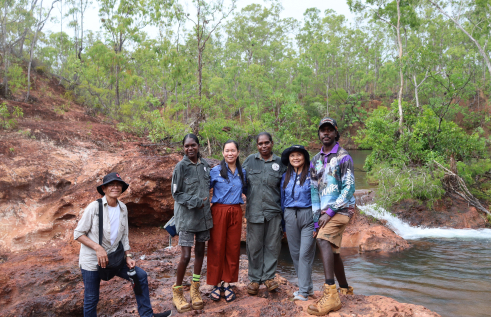 Six people looking at camera smiling standing on a rock in front of a waterfall.
