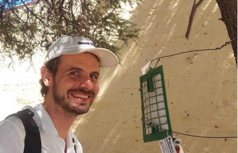 Andrew Coates, head and chest, on the left of the photo, in a white shirt and a white cap, smiling at the camera, in front of a white wall, with tree branches above his head. A bug trap attached by wires to a tree trunk which is just visible at the right of the image