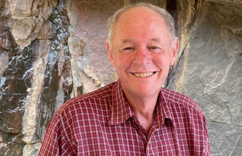 Steven Tickell, head and chest, wearing a red shirt with white cross hatching, with a rock face behind him